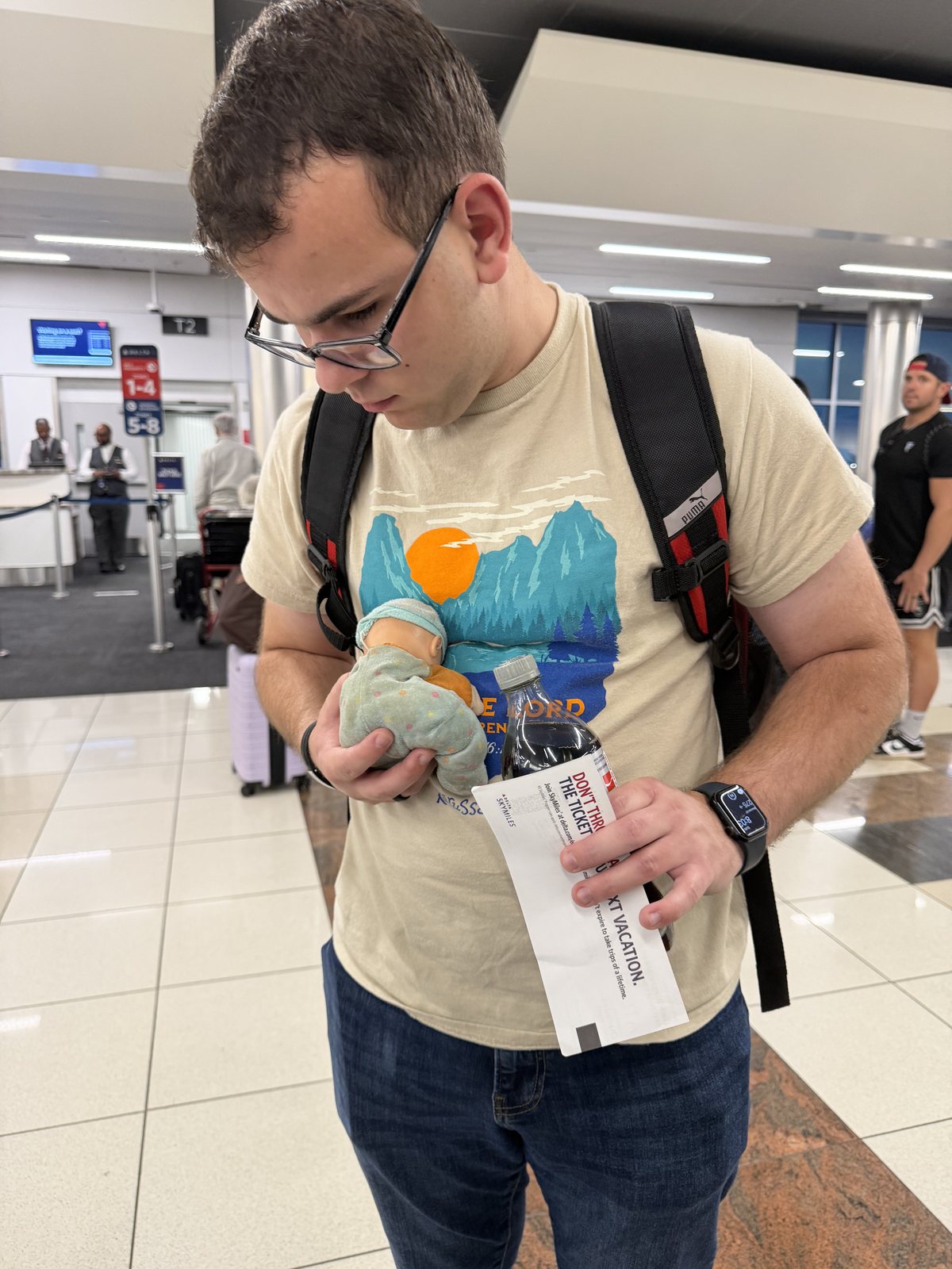 ATL. Liem with his boarding pass in one hand and Lucy — Travis's doll — in the other, holding her for him.